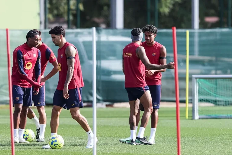 Jogadores do FC Barcelona de vermelho e azul-marinho num treino no relvado, com balizas e um cone ao fundo.