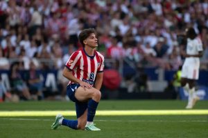 Football player in red and white striped kit with Atlético Madrid crest, kneeling on the pitch in a stadium before a crowd.