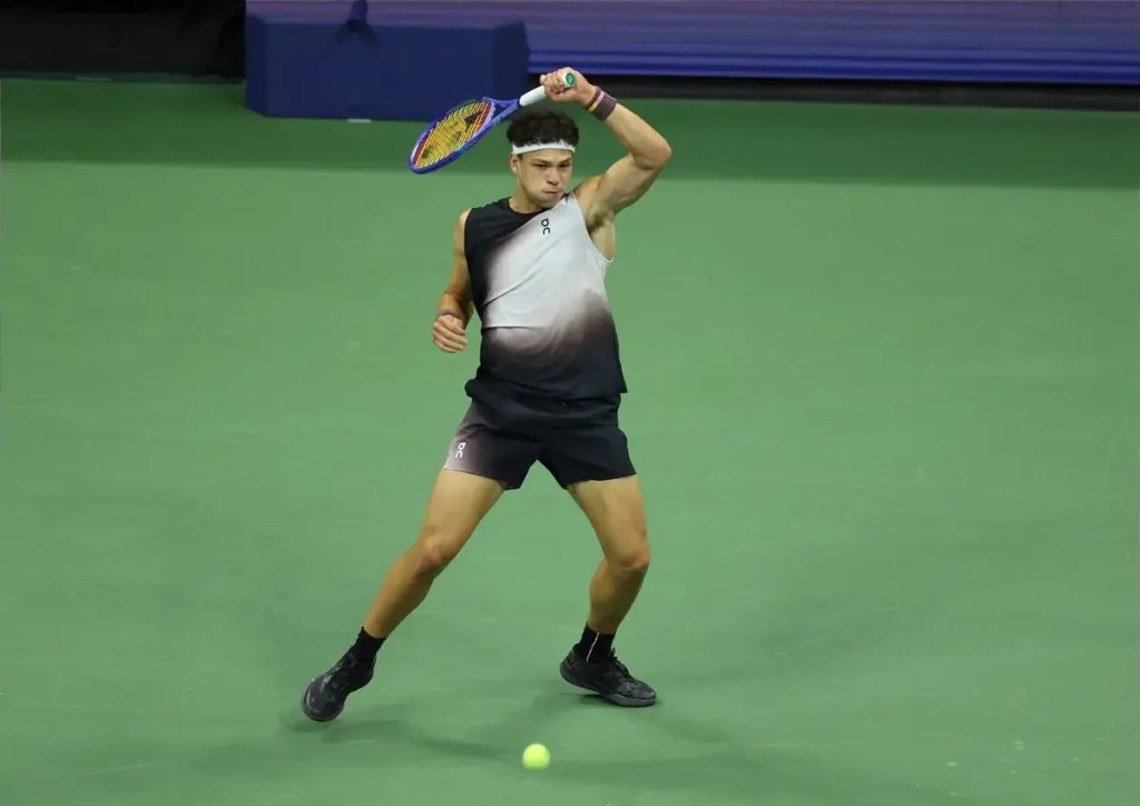 Tennis player in a black-and-grey sleeveless top and dark shorts hitting a forehand on a green court, racket raised, ball near his feet. Ben Shelton, per the…