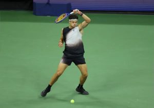 Tennis player in a black-and-grey sleeveless top and dark shorts hitting a forehand on a green court, racket raised, ball near his feet. Ben Shelton, per the…