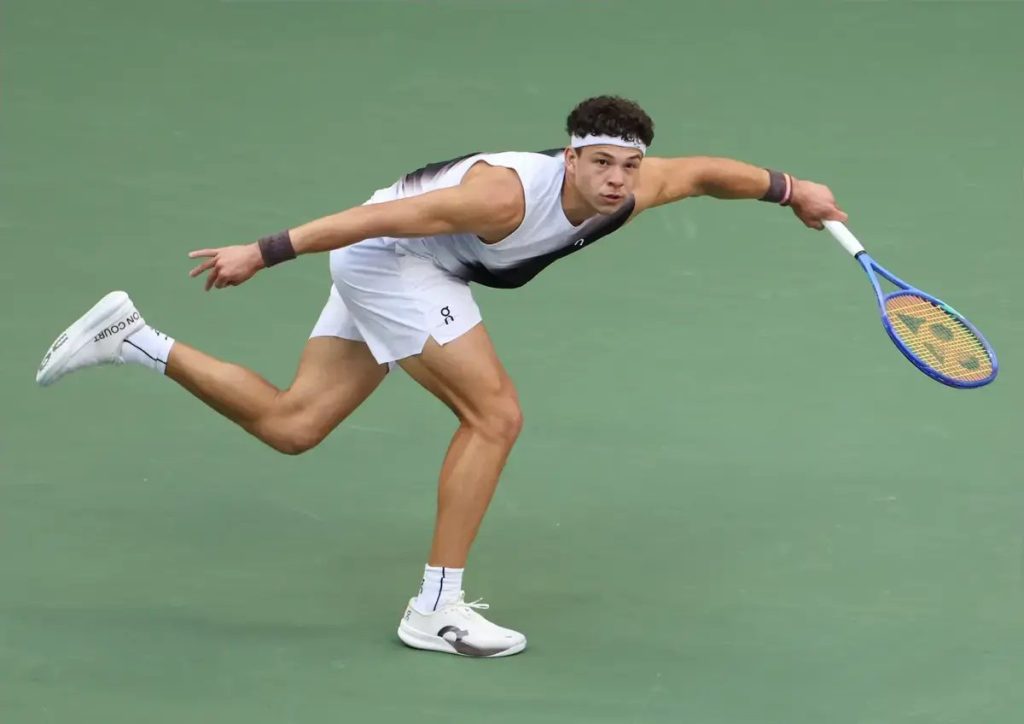 Tennis player in white outfit and headband stretching for a wide shot, swinging a blue Yonex racket on a green court