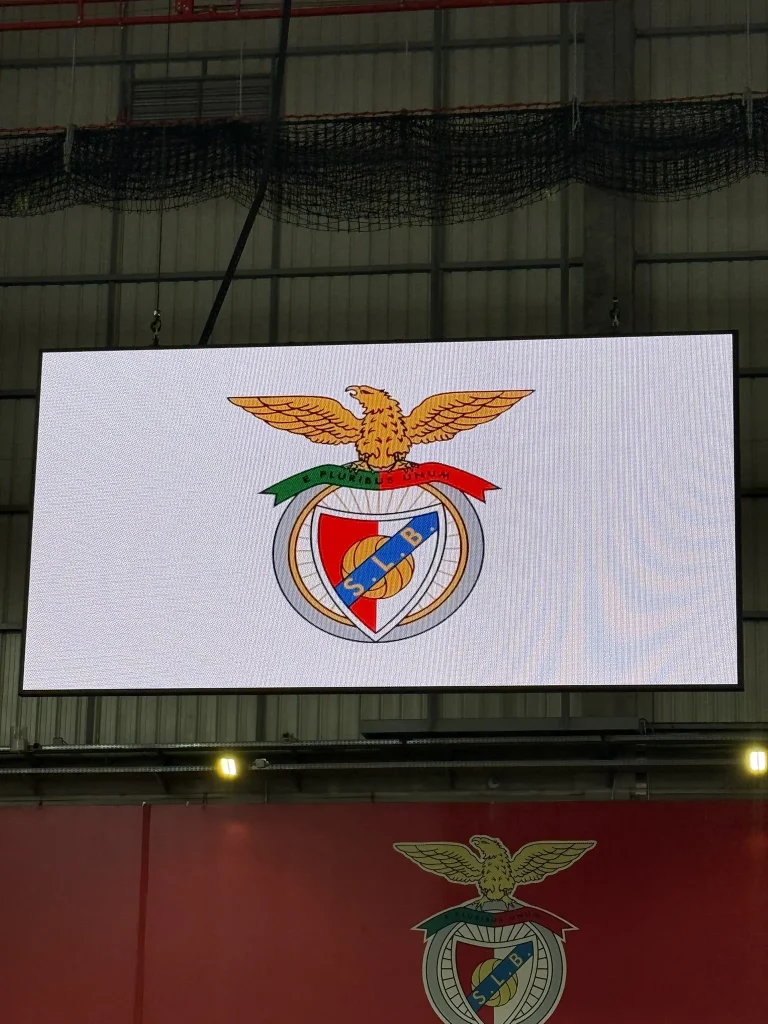 Ecrã gigante no Estádio da Luz com o emblema do Sport Lisboa e Benfica, águia dourada sobre escudo vermelho e azul com as iniciais SLB.
