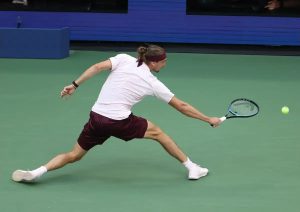 Tennis player in white shirt and maroon shorts stretching low to return a ball with a blue racket on a green court
