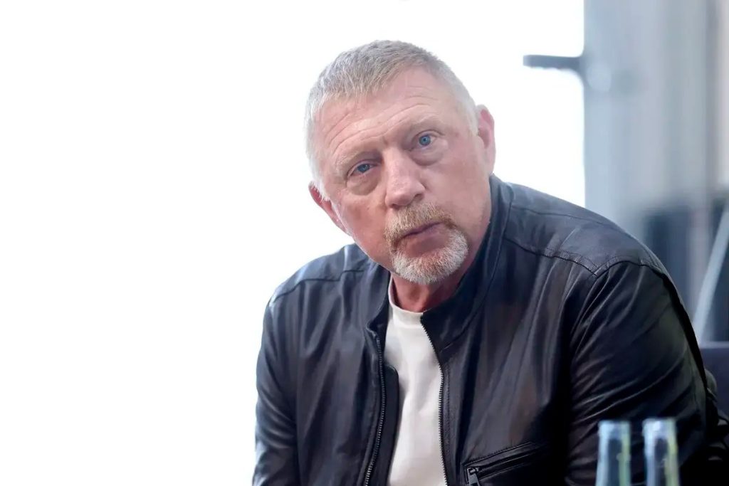 A middle-aged man with short gray hair and a gray beard, wearing a black leather jacket over a white shirt, speaking in an indoor setting with a bright backg…