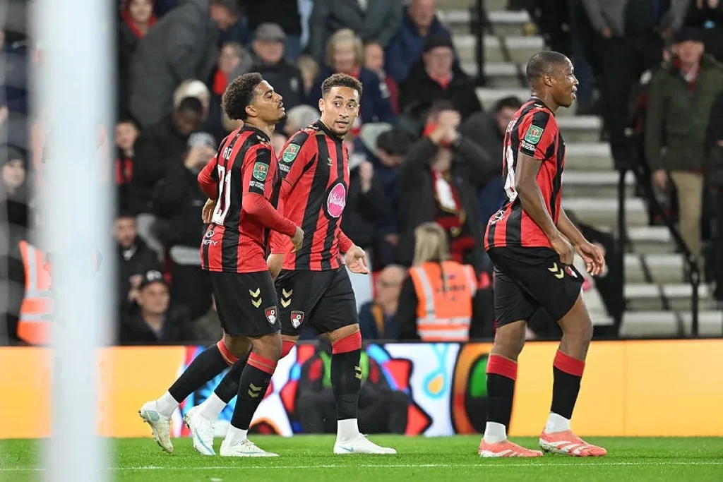 Três jogadores do Bournemouth, de listas vermelhas e pretas, celebram ou caminham em campo, com adeptos ao fundo num estádio.