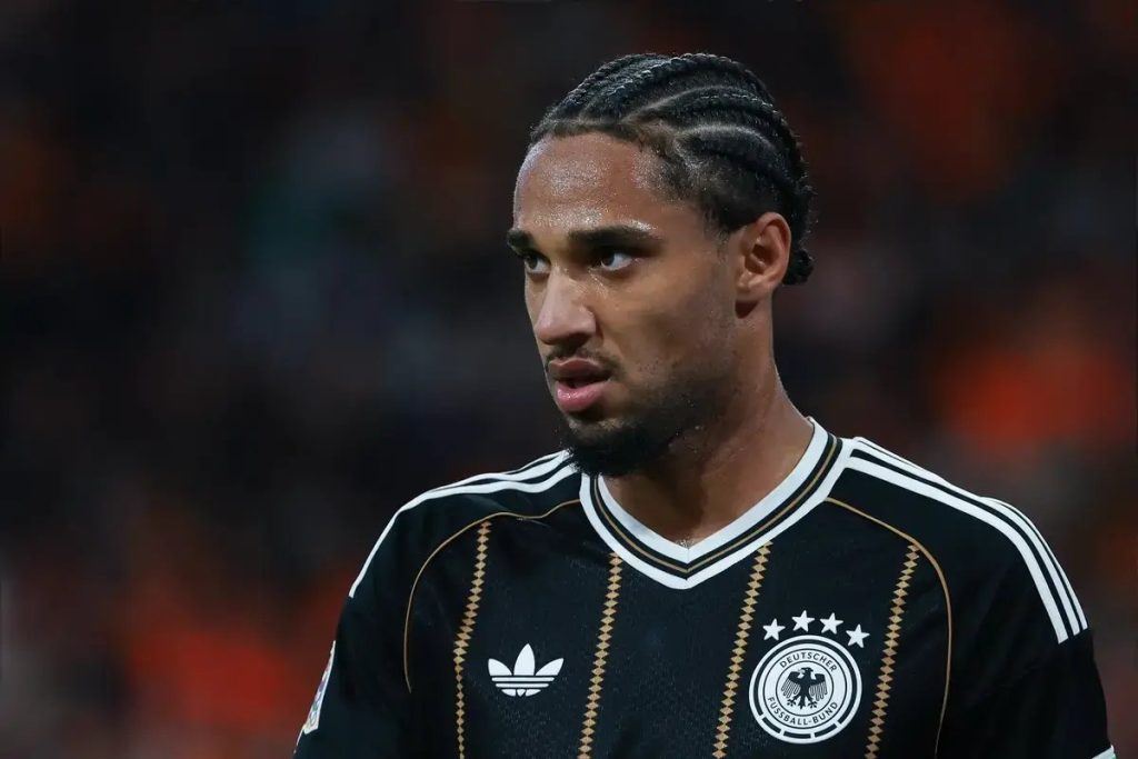 Football player in a black Germany national team shirt with braided hair, looking to the side at a match. Crowd blurred in background.
