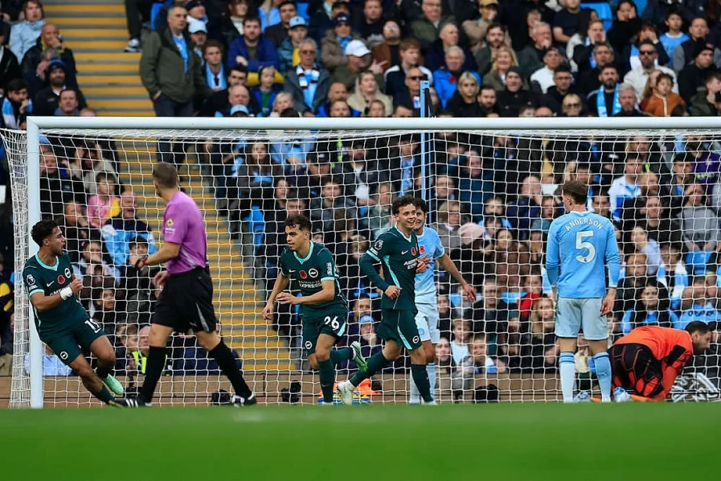 Jogadores do Brighton de verde celebram um golo no Etihad, enquanto um defesa do Man City de azul claro, nº 5, observa o lance.
