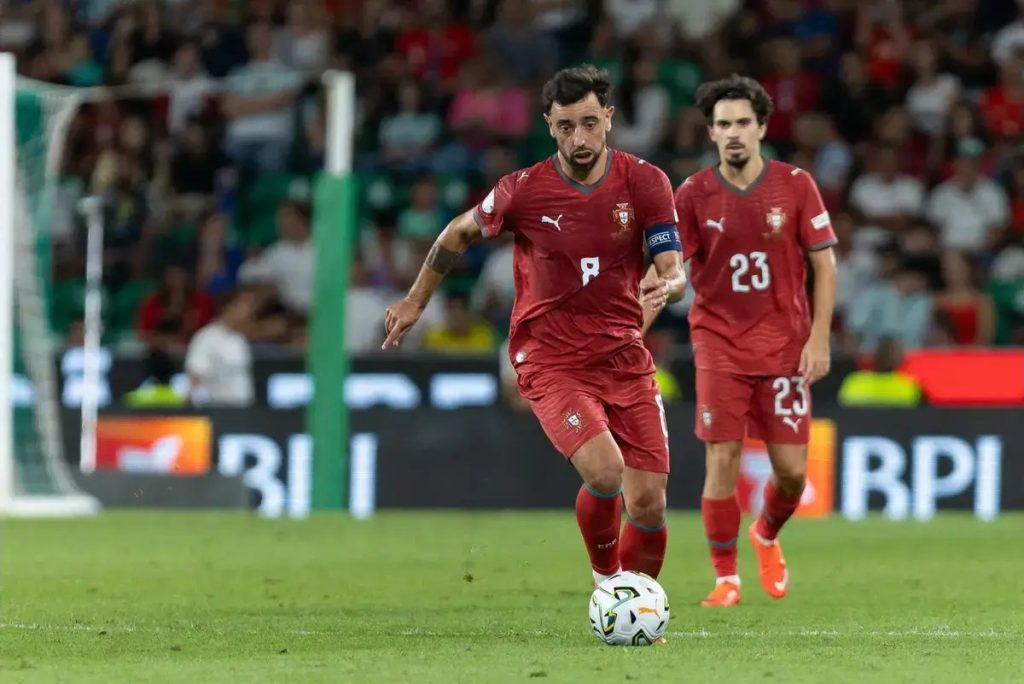 Player #8 in red Portugal kit with captain's armband dribbles the ball on the pitch, teammate #23 behind him, stadium crowd in background