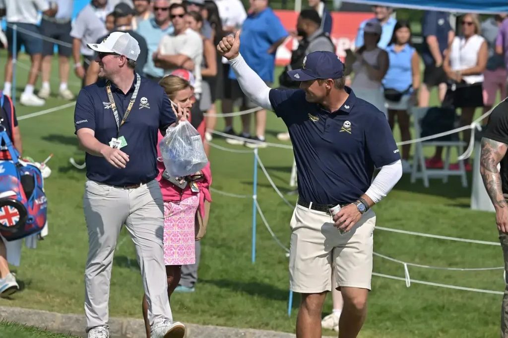 Man in navy polo with skull-and-crossbones logo gives a thumbs-up while walking a golf course lined with spectators and ropes