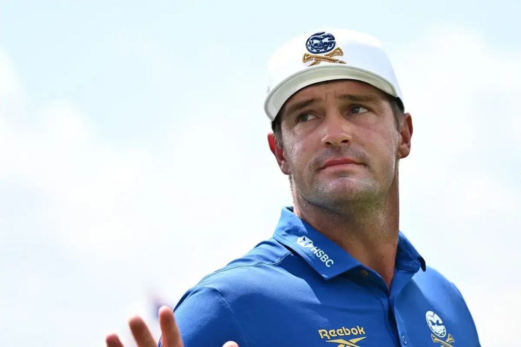 Golfer in a white cap with a blue polo bearing HSBC and Reebok logos, gesturing with an open hand on a golf course.