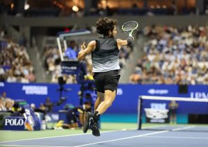 Tennis player mid-jump celebrating on court, holding a racket, at a blue-court arena with American Express and Rolex signage; name not visible in photo.