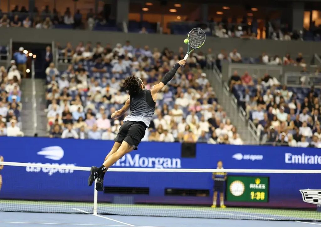 Tennis player in black outfit leaping to strike a forehand overhead at the US Open, with the net and packed stands behind him.