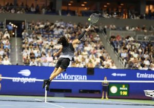 Tennis player in black outfit leaping to strike a forehand overhead at the US Open, with the net and packed stands behind him.
