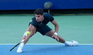 Tennis player in dark Nike kit stretches low to reach a yellow ball with his backhand on a blue hard court.