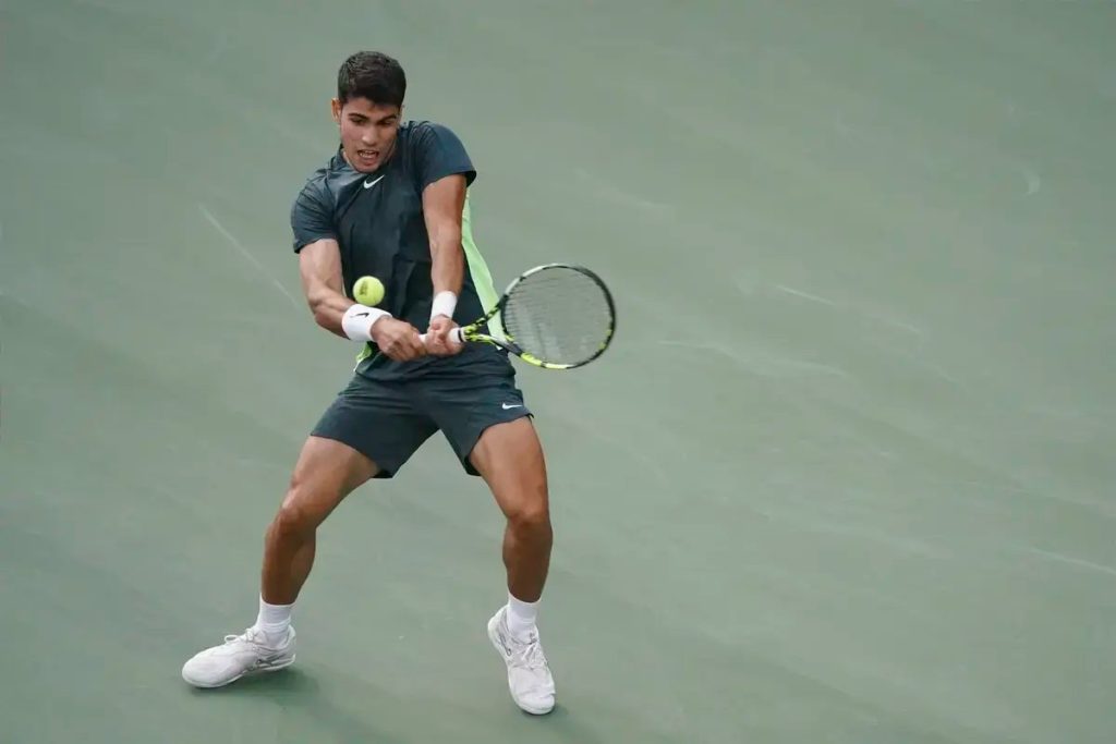 Tennis player in dark green Nike outfit and white wristband hits a backhand on a green hard court, eyes on the ball