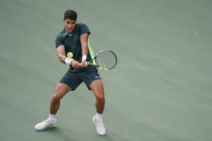 Tennis player in dark green Nike outfit and white wristband hits a backhand on a green hard court, eyes on the ball