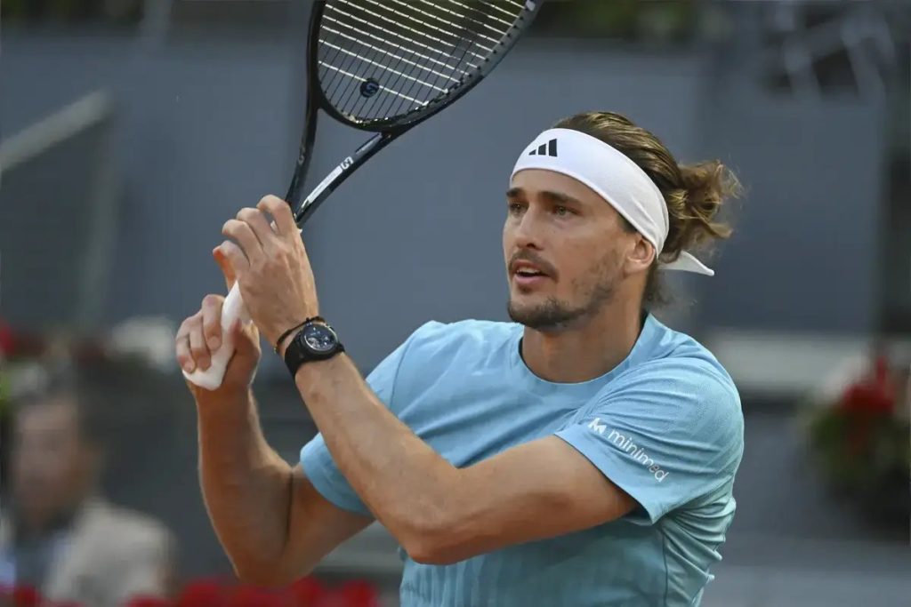 Tennis player in a light blue shirt and white Adidas headband, gripping his racket mid-swing on a clay court in a match setting.