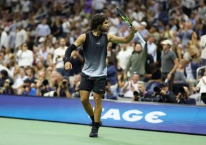 Tennis player in a black sleeveless top and dark shorts, raising his racket to fans at a US Open match, with photographers crowded behind the court.