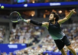 A tennis player in a black sleeveless shirt and black shorts, arm outstretched to return a yellow ball at a US Open match.
