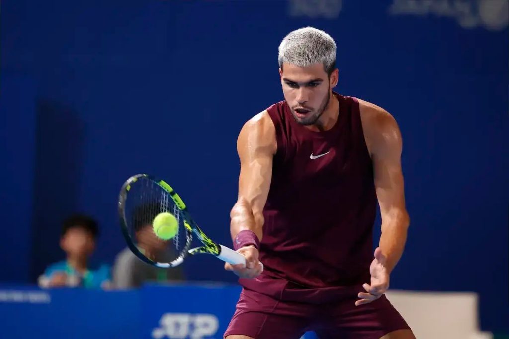 Tennis player with bleached spiky hair in a maroon Nike sleeveless top hits a low forehand on a blue court, tracking the neon-green ball.