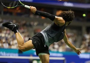 Tennis player in black sleeve, black shorts and grey tank top, mid-stroke with a Babolat racket at a night match, arm and leg stretched out.