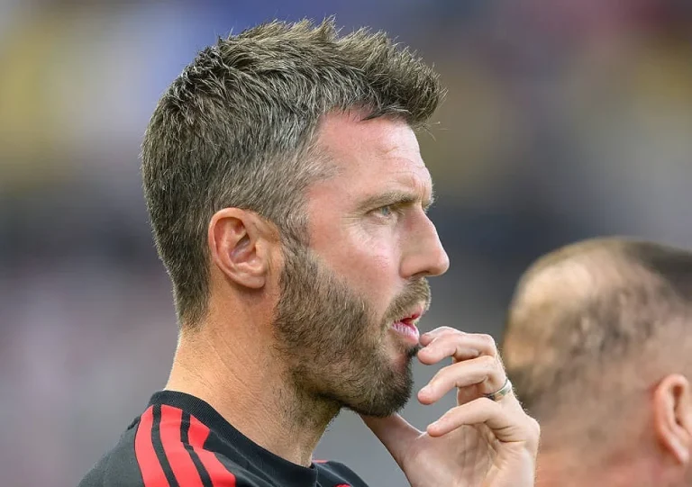 Homem de cabelo curto e barba, de perfil, com camisola preta e riscas vermelhas, a levar a mão ao queixo junto à linha lateral de um estádio.
