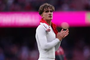 Young footballer with curly hair in white kit and red scarf, hands pressed together, applauding fans in a stadium. Article about Max Dowman transfer talks.