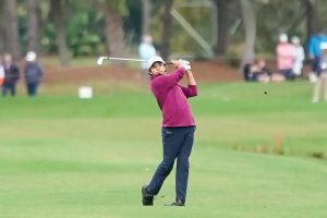 Golfer in a maroon top and navy trousers takes a fairway iron shot on a green course, with spectators watching behind him.