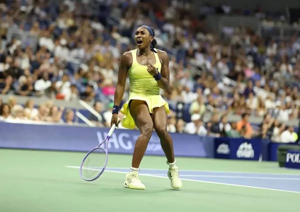 Coco Gauff celebrates fiercely on court in a yellow dress and headband during a tennis match at a packed stadium.