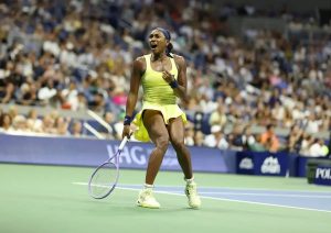 Coco Gauff celebrates fiercely on court in a yellow dress and headband during a tennis match at a packed stadium.