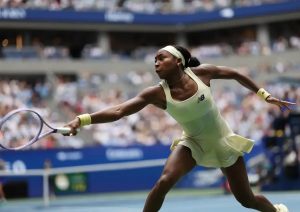 Tennis player in a pale yellow New Balance dress and headband, stretching to reach a backhand during a match on a blue court.