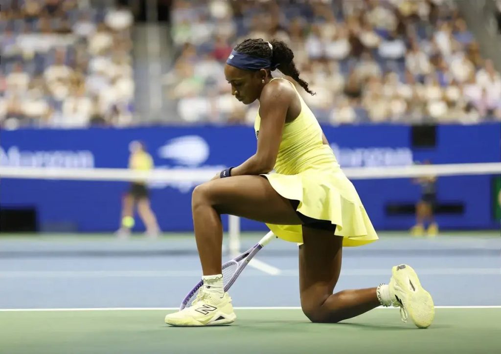 Tennis player in yellow dress and navy headband kneeling on court, head bowed, racket beside her, crowd blurred behind