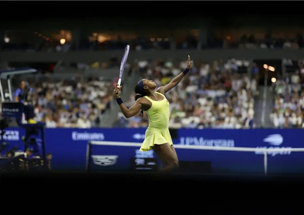 Tennis player in a yellow dress and navy headband raises her racket and arms in celebration on a packed stadium court.