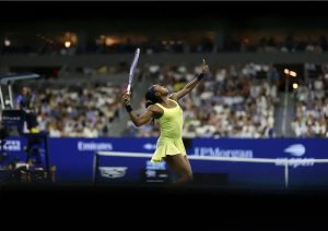 Tennis player in a yellow dress and navy headband raises her racket and arms in celebration on a packed stadium court.
