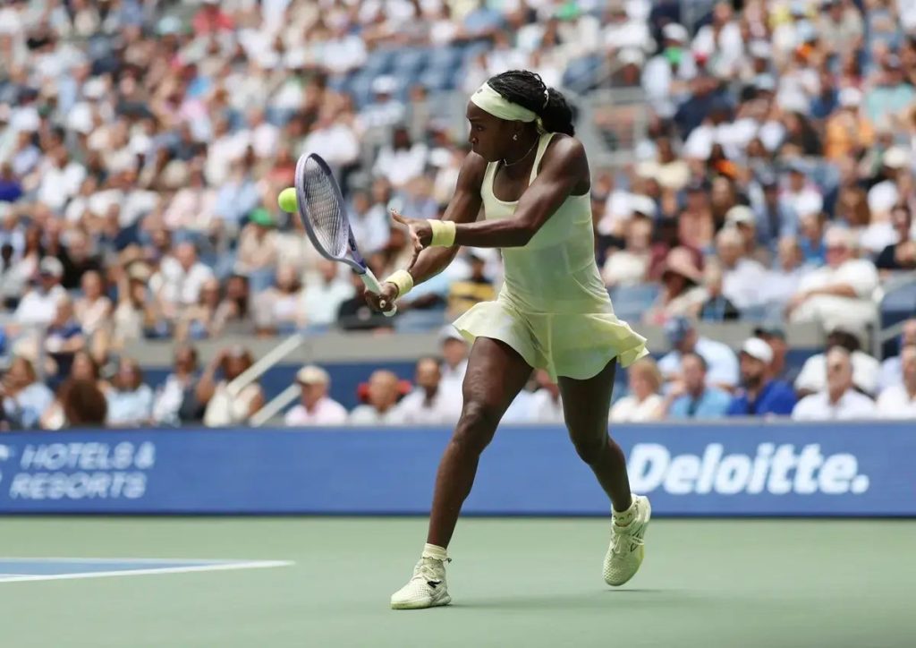A tennis player in a pale yellow dress hits a forehand during a match at a packed stadium, with a Deloitte sign behind her.