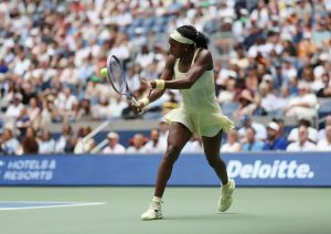 A tennis player in a pale yellow dress hits a forehand during a match at a packed stadium, with a Deloitte sign behind her.