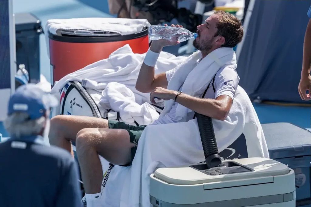 Tennis player in white shirt and towel drinking water from a bottle while seated on a courtside cooler at a hard court event