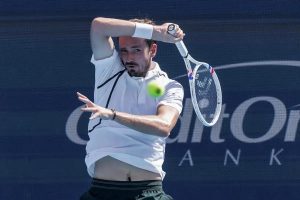 Tennis player in white polo and dark shorts hits a forehand near a yellow ball, racket raised, blue Citi Open backdrop