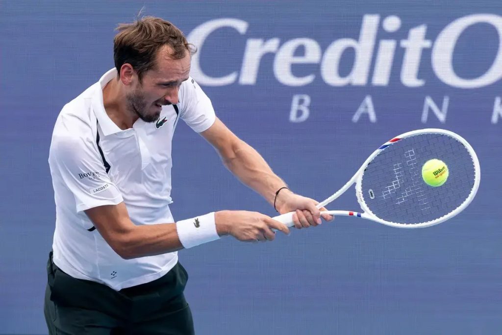 Tennis player in white Lacoste polo and dark shorts hits a backhand with a Wilson racquet, a yellow ball in view, against a Credit One Bank backdrop.
