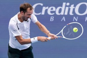 Tennis player in white Lacoste polo and dark shorts hits a backhand with a Wilson racquet, a yellow ball in view, against a Credit One Bank backdrop.