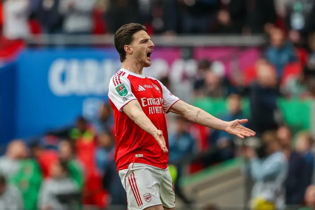 Footballer in red and white Arsenal kit with Emirates sponsor, arms outstretched and shouting on the pitch at a match
