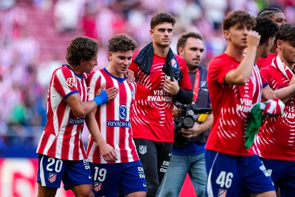 Atlético Madrid players in red-and-white stripes, numbers 20 and 19, embrace after a match as a teammate in a red training top walks past a photographer.