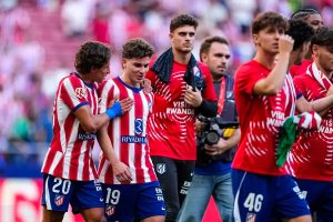 Atlético Madrid players in red-and-white stripes, numbers 20 and 19, embrace after a match as a teammate in a red training top walks past a photographer.