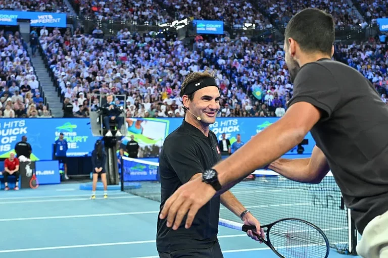 Dois tenistas sorriem num court azul durante um evento com público, um de costas e outro de t-shirt preta e faixa na cabeça, com um cartaz de patrocinador ao…