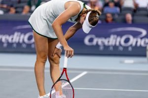 Tennis player in white visor and dress, bent over clutching her leg in pain on a court at a CreditOne event.