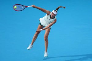 Tennis player in white Yonex outfit and headband serving on a blue court, racket extended, ball visible in the air