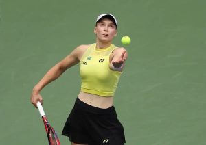 A tennis player in a yellow Yonex top and black skirt tosses a ball overhead to serve on a green hard court
