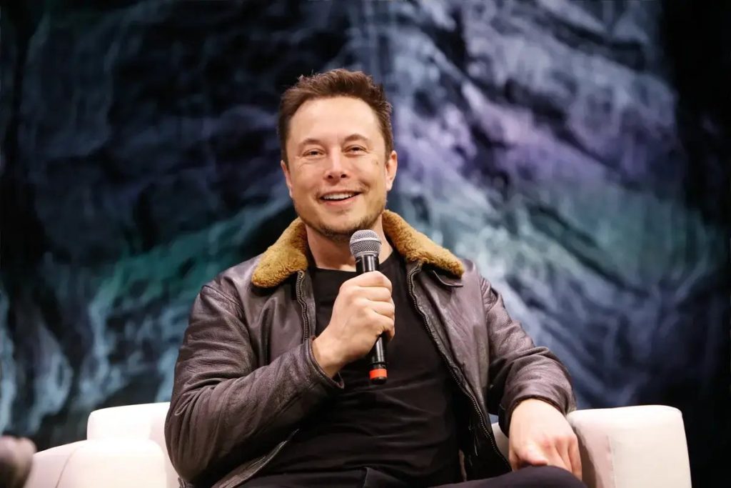 A smiling man in a brown shearling-collar leather jacket holds a microphone while seated on a white chair in front of a dark, blue-green backdrop.