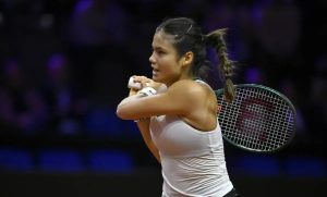 A tennis player in a white top and ponytail, holding a Wilson racket, mid-swing on a dark indoor court at a night match.