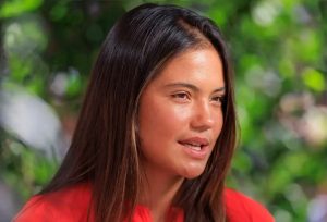 Woman with long dark hair in a red top, speaking outdoors against a blurred green background, appearing in an interview setting